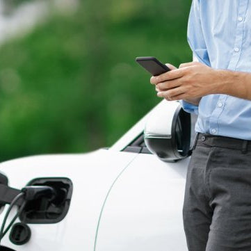 A man checking his smartphone while leaning against his white charging EV.