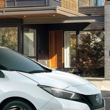 A white Nissan Leaf electric car parked in front of a modern, multi-story house with a brick facade and a black garage door.