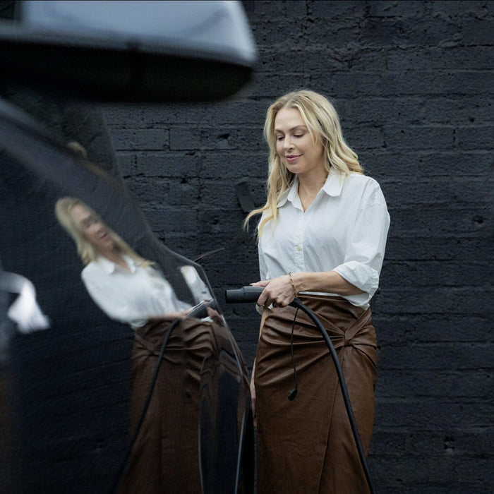A blonde woman in a white blouse and brown skirt holds a charging handle to plug into a black EV while standing next to a wall-mounted EV charger on a dark brick wall.