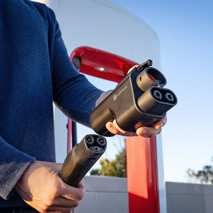 A person holding a slim NACS charging handle in one hand and a bulky black Lectron NACS-to-CCS1 adapter in the other at a charging station.