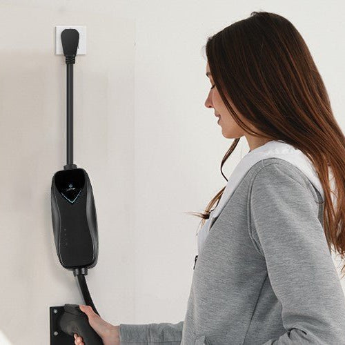 A woman in a grey hoodie standing next to a wall-mounted Lectron Level 1 EV charger plugged into an indoor outlet.