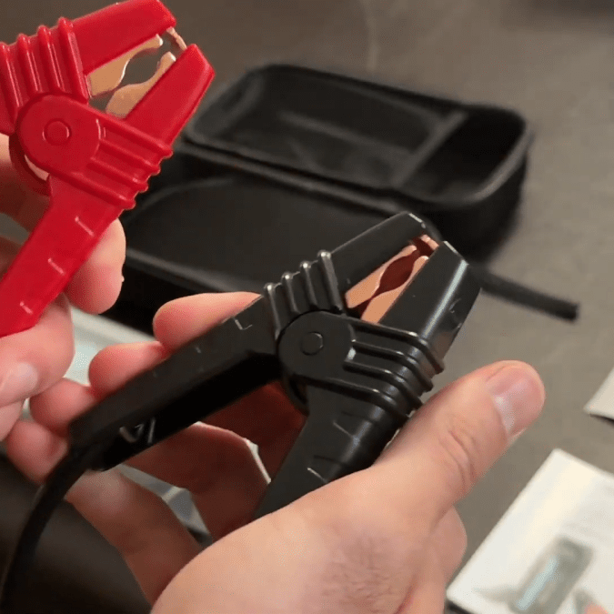 A person holding a pair of heavy-duty jumper clamps, one red and one black.