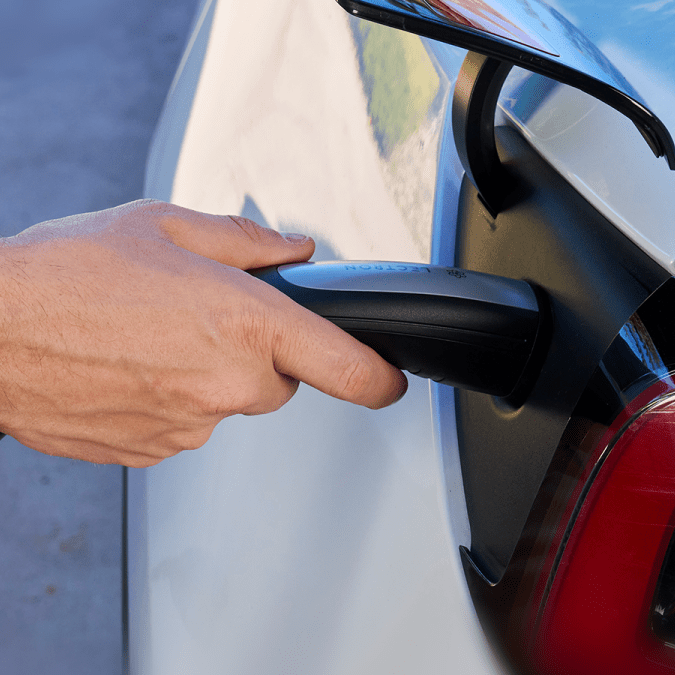 A person's hand plugging a black Lectron EV charging handle into the charging port of a white Tesla vehicle.