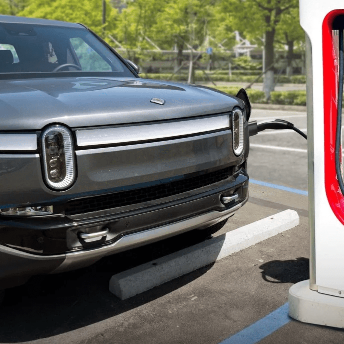 A grey Rivian R1T electric pickup truck plugged into a white and red Tesla Supercharger station in a parking lot.