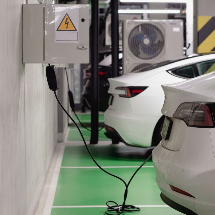 An indoor garage charging bay featuring white Tesla vehicles, where a black cable connects a wall-mounted power box to the rear charging port of the closest car