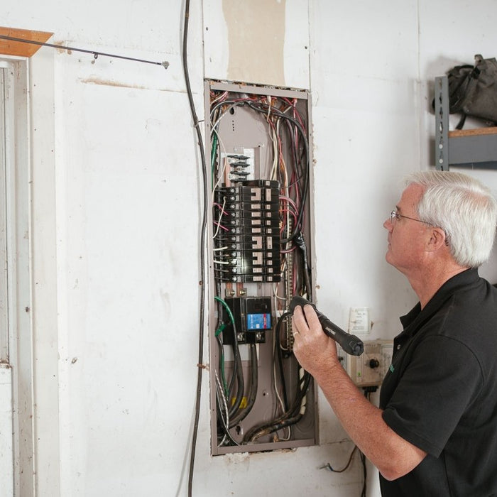 An electrician inspecting an exposed electrical breaker panel box with a flashlight inside a home or garage.