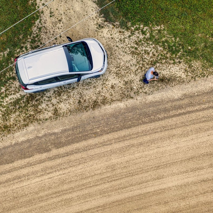 Aerial top-down view of a white hatchback car parked alongside a dirt road next to a person standing in a grassy field.