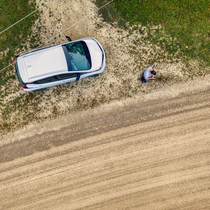 Aerial top-down view of a white electric crossover vehicle parked on a grassy field edge next to a dirt road.